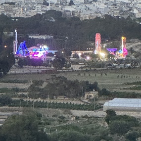 Distant nighttime view of the Magic World amusement park in Malta featuring illuminated carnival rides and city lights.
